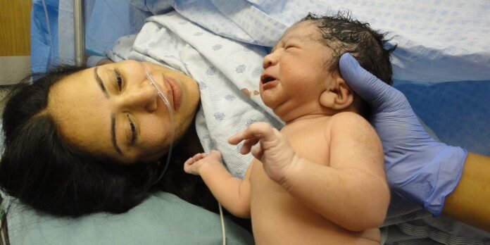 Mother lying in a hospital bed with an oxygen nasal cannula, gazing at her newborn baby supported by a gloved caregiver's hand.