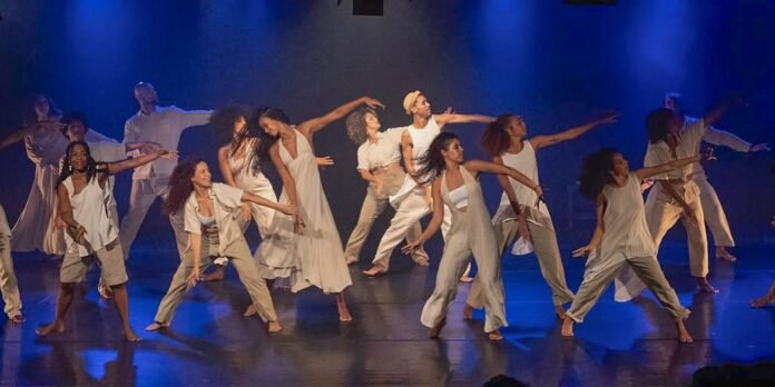 Group of diverse dancers in beige outfits performing a choreographed routine on a blue-lit stage, barefoot and expressive.