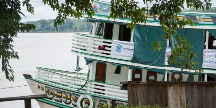 Green-and-white two-story riverboat named 'DEUS' docked along a muddy river, banners on the upper deck and leafy branches in the foreground.