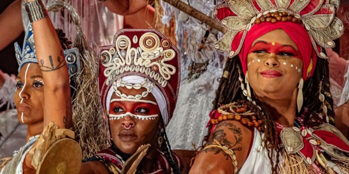 Group of carnival dancers in colorful costumes; central figure wears a shell crown and red face paint, flanked by two others with face paint and beads, smiling.
