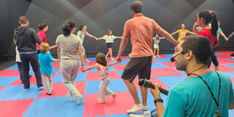 Musician in a teal shirt with a headset plays guitar as a diverse group of kids and adults form a circle, holding hands on a red-and-blue gym mat.