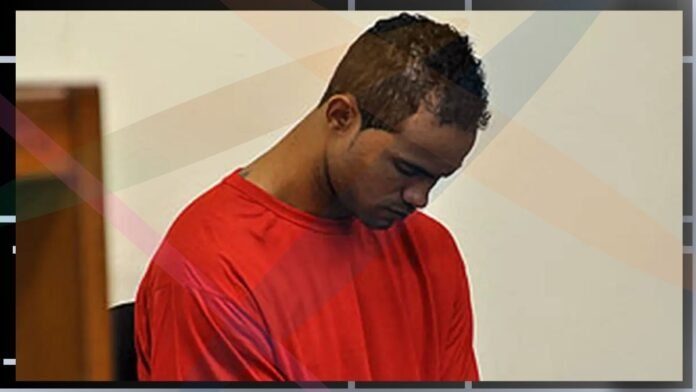 Man in a red shirt looking down while seated indoors, neutral setting.