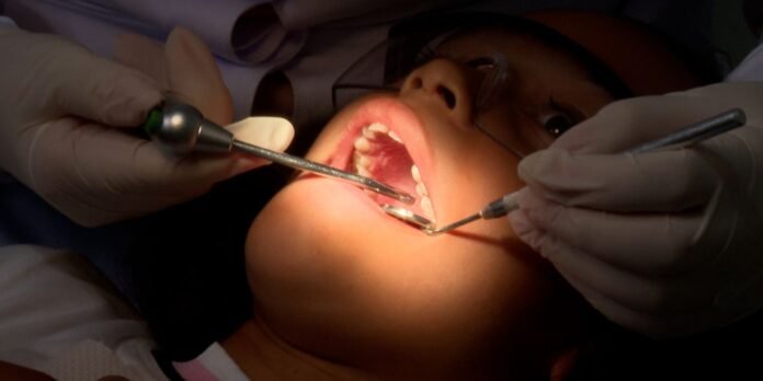 Close-up of a dental procedure: gloved hands hold dental instruments inside a patient's open mouth.