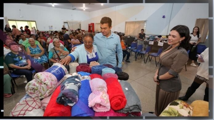 A man helps an elderly woman choose colorful blankets at a crowded community event in a hall for a blanket donation drive.