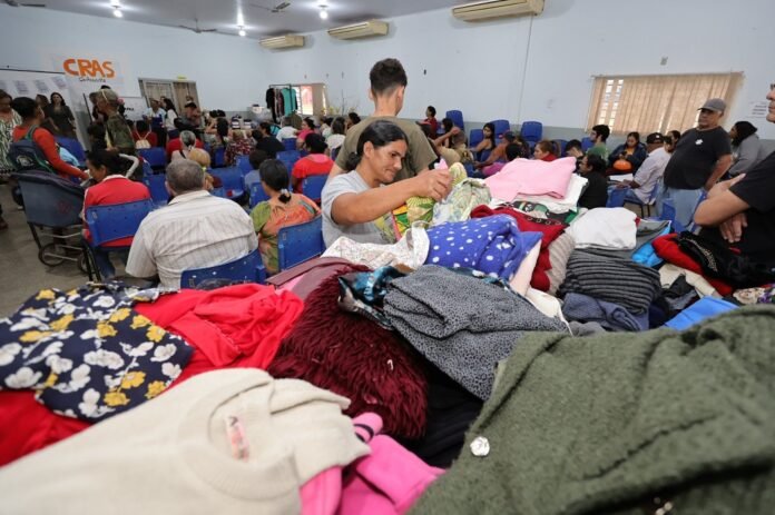 Woman sorting through a pile of donated clothes at a crowded relief center with people seated in blue chairs in the background.