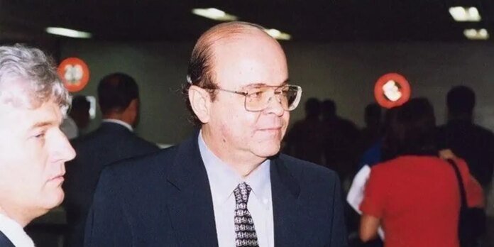Man in a dark suit and patterned tie walking through a crowded indoor event, seen in profile with other attendees in the background.
