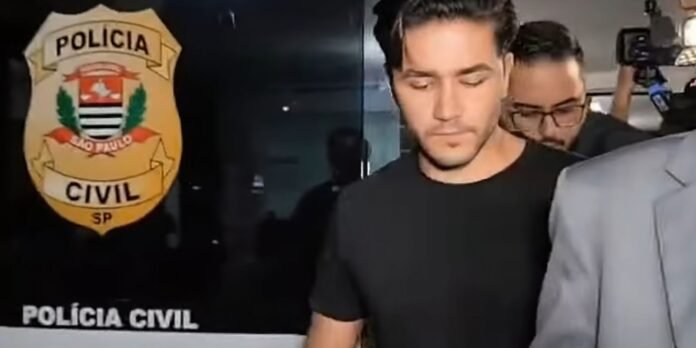 Man in a black T-shirt escorted by authorities, with a Civil Police badge visible in the background and media filming nearby.