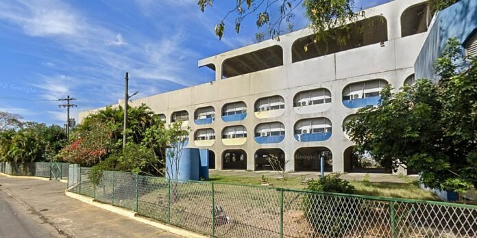 Concrete multi-level parking garage with rounded openings and blue accents, bordered by a green chain-link fence and tropical plants under a blue sky.