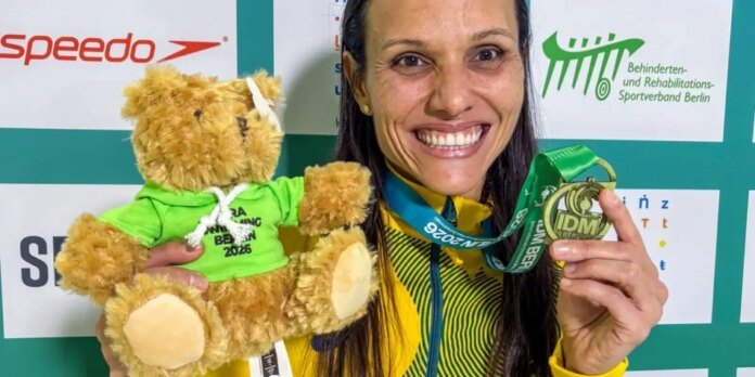 Smiling female athlete holding a gold medal and a plush teddy bear in a green hoodie, with sponsor banners in the background