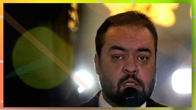Close-up of a man with dark hair and beard speaking into a microphone on a dim stage with bright lights behind him.