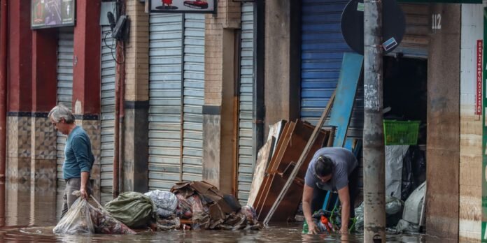 Older man wading through floodwater, lifting bags of trash along a storefront-lined street with closed metal shutters and debris in the water.