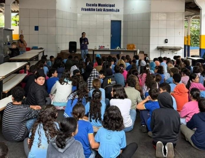 School children sit cross‑legged on the floor in a covered hall, facing a speaker at a front stage; a sign reads 'Escola Municipal Franklin Luiz Azambuja'.