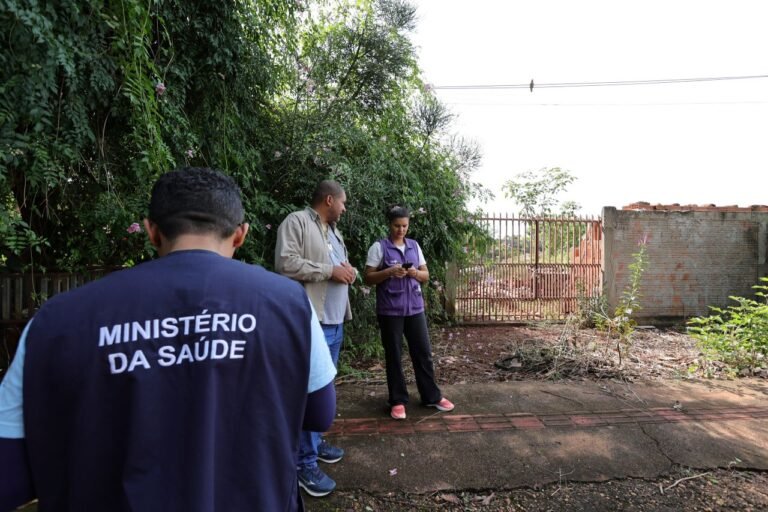 People standing outdoors near a green hedge; one person wears a vest reading 'MINISTÉRIO DA SAÚDE' as they look at a phone.