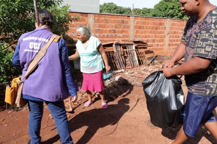 People cleaning up a rubble-filled yard: a woman in a teal top and pink skirt talks to another person while a man walks by carrying a black trash bag; piles of bricks and debris against a brick wall are in the background.