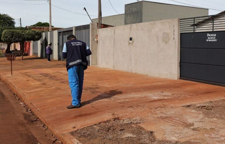 Health Ministry worker in blue pants and a dark vest labeled 'MINISTÉRIO DA SAÚDE' walks along a dusty street beside concrete walls and a large black gate.