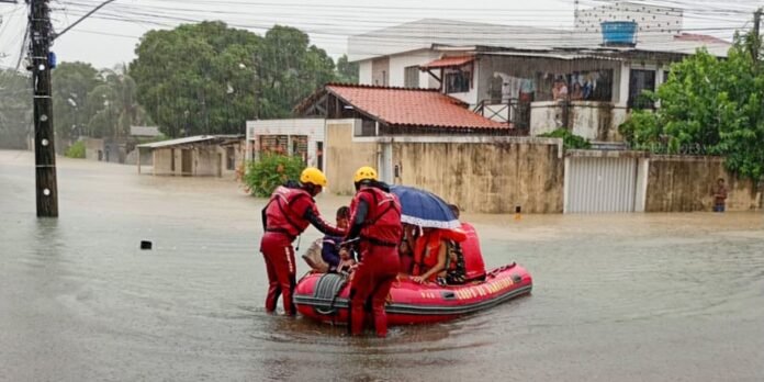 Rescue workers in red suits and yellow helmets evacuate people in a red inflatable boat through a flooded street.
