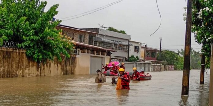 Rescue workers in yellow helmets and red gear navigate waist-deep floodwaters with inflatable boats in a flooded residential street, a dog nearby.
