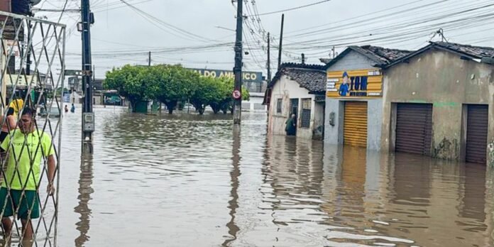 Urban street severely flooded, water halfway up storefronts and signs, pedestrians wading through murky water with utility poles overhead in a residential-commercial block (likely after heavy rain).