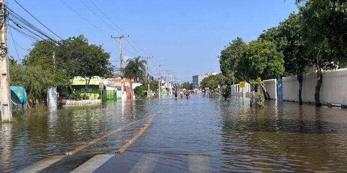 Flooded urban street with water covering the road; people wade in the distance beside trees and buildings on both sides.