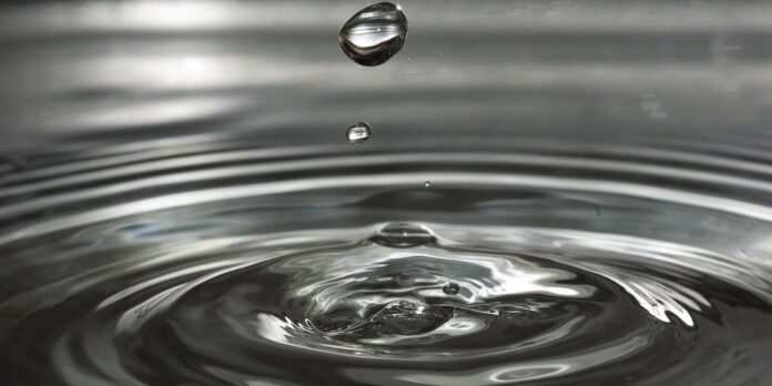 Macro photo of a water droplet just above a rippling water surface, with concentric waves spreading outward.