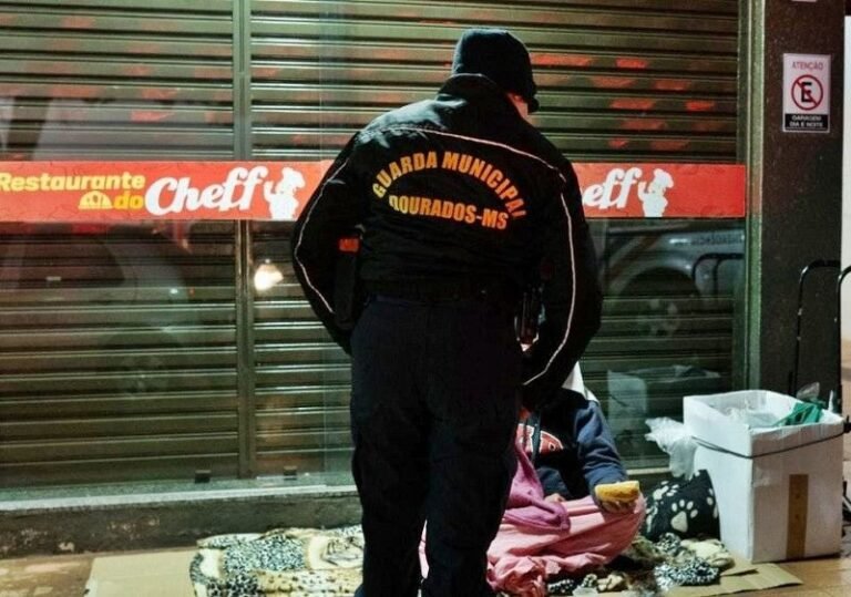 Municipal guard in uniform standing over a person sitting on the ground with blankets near a restaurant storefront at night.