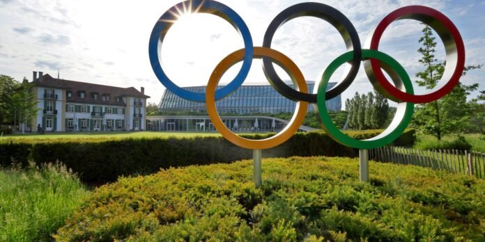 Colorful Olympic rings sculpture in a park with a modern glass building in the background, sunlight streaming through the rings.