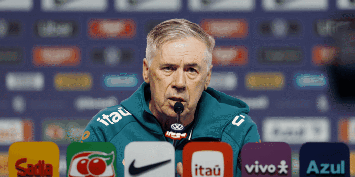 Older man in a teal jacket speaks into a microphone at a press conference with sponsor logos blurred in the background.