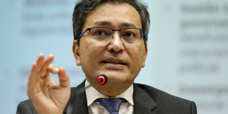 Man in a suit and tie speaks into a red-ringed microphone, gesturing with his hand while wearing glasses in a conference setting.