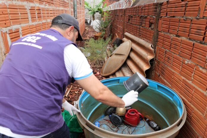 Worker in a purple vest and white gloves handles a metal component over a large blue plastic tub filled with pump parts in a narrow outdoor alley between red brick walls with barbed wire on top.