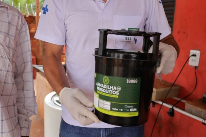 Person wearing gloves holds a black container labeled 'Armadilha Mosquitos' (mosquito trap) for pest control in a workshop setting