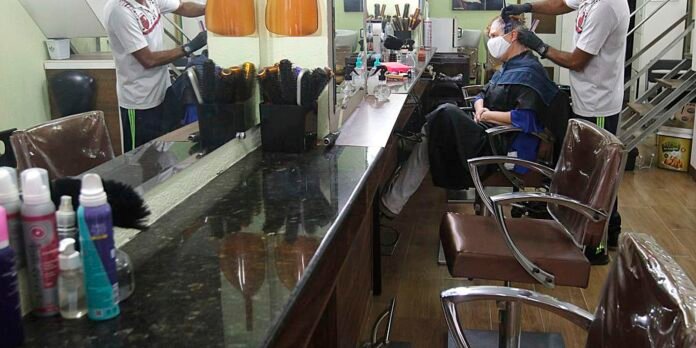 Barber shop scene: client wearing a mask sits in a chair while a barber trims hair, another stylist stands at the mirror, and hair products line the counter behind them.