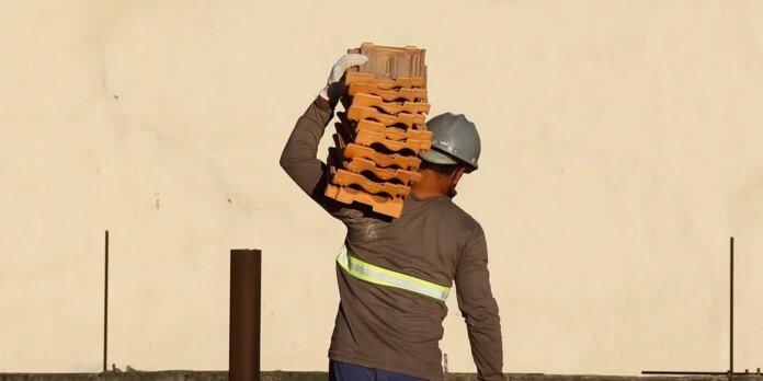 Construction worker wearing a hard hat and reflective vest carries a tall stack of red bricks on his shoulder outdoors against a beige wall.