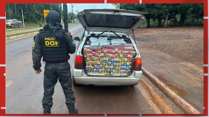 Officer in tactical gear stands beside a white car with its trunk open, revealing hundreds of boxed cigarettes stacked inside.