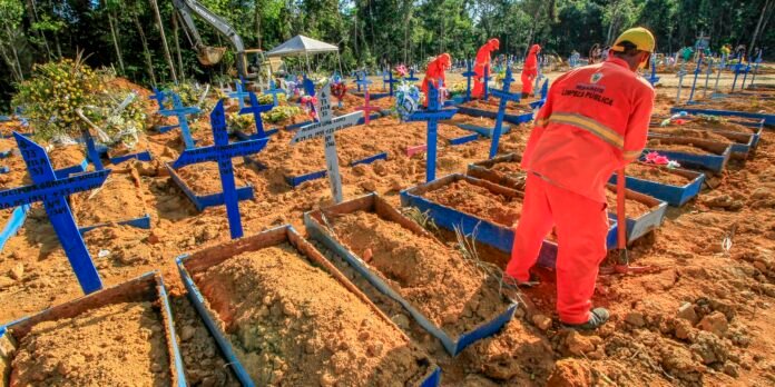 Workers in bright orange uniforms dig graves in a cemetery with blue wooden crosses and orange soil, greenery in the background.