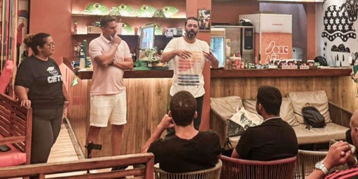 Three men and a woman stand at a casual presentation area in a tropical-themed lounge, facing a small seated audience by a wooden bar.