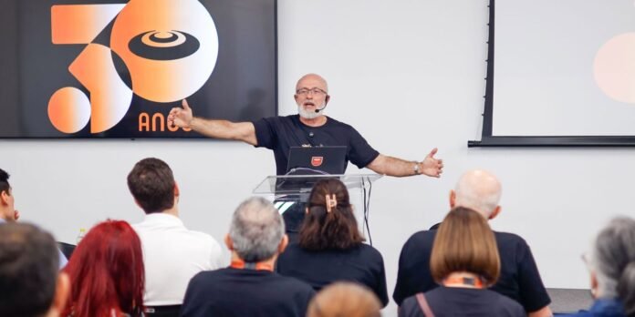 Speaker with headset mic presenting to an audience in a conference room, gesturing with open arms behind a clear lectern.