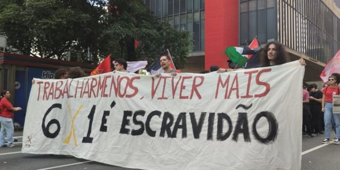 Protesters on a city street hold a large white banner with red text and bold black lettering, signaling a workers' rights demonstration.
