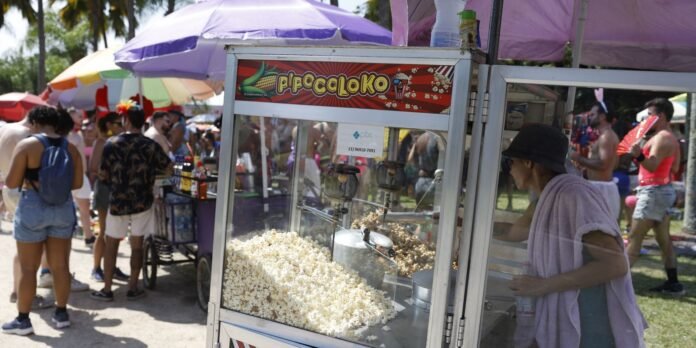 Popcorn cart at a sunny outdoor festival with a glass display and a vendor wearing a hat, crowd and colorful umbrellas in the background.