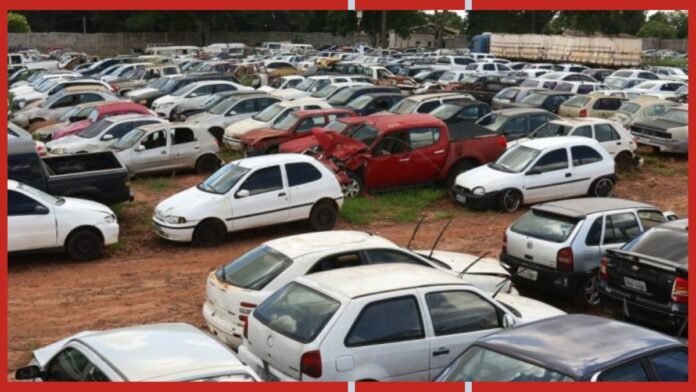Crowded junkyard filled with old, damaged cars parked on dirt ground adjacent to each other, under a fence-bound lot.