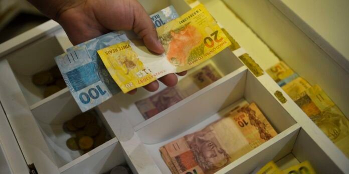 Hand placing colorful banknotes into a cash register drawer with coins visible below.