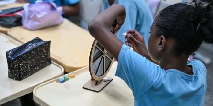 Student in a blue shirt working on a small wooden wheel at a classroom craft station, with paint bottles nearby