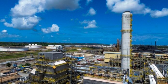 Industrial refinery with a tall smokestack, yellow safety railings, and extensive pipelines under a blue sky.