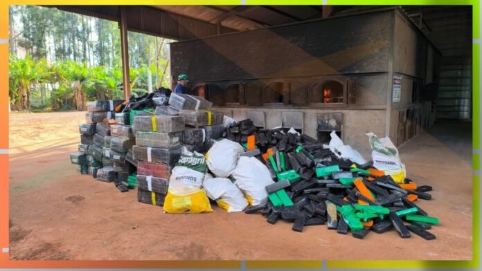 Piles of black and green plastic blocks and white bags stacked outside a large industrial kiln; a worker in a hard hat nearby under a covered area.