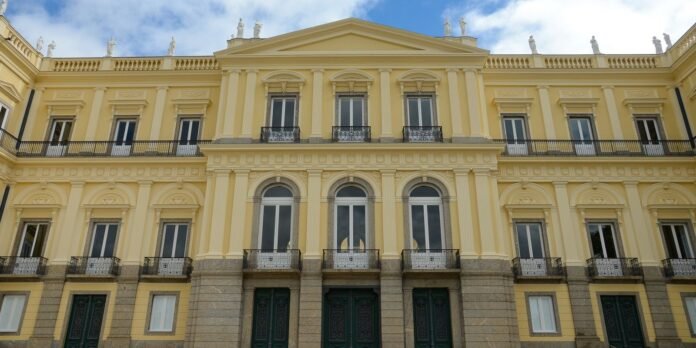 Grand pale-yellow neoclassical building facade with arched windows, decorative columns, and wrought-iron balconies, set against a blue sky with statues atop the roofline.