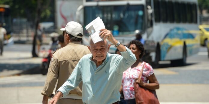 Elderly man in a light blue shirt holds a folded newspaper to his head on a sunny city street; a bus and pedestrians are in the background.
