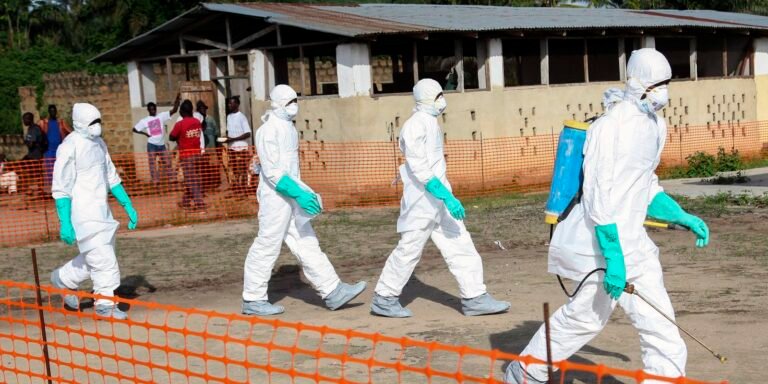 Four people in white hazmat suits with teal gloves walk in a cordoned-off area, one carrying a spraying device on their back and a wand in hand.