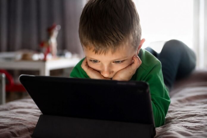 Young boy in a green shirt lies on a bed, propped on his elbows, focused on a tablet in a black case at home.