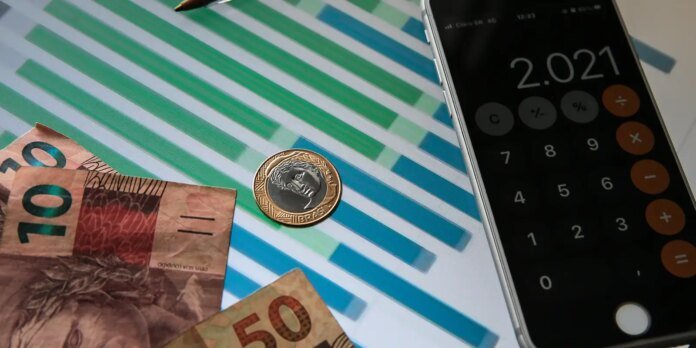 Brazilian reais banknotes and a 1 real coin beside a smartphone calculator showing 2.021, on a striped background, suggesting budgeting or calculation.