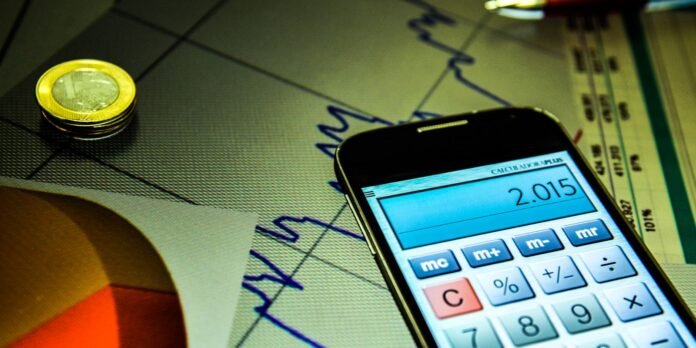Smartphone showing a calculator app with 2.015 on the screen, beside a coin and financial documents on a desk.