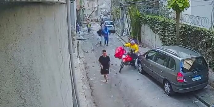 Narrow urban alley with a parked gray car on the right; a man in black clothing walks toward the camera while a motorcyclist in yellow sits nearby and debris is scattered on the street in the background.